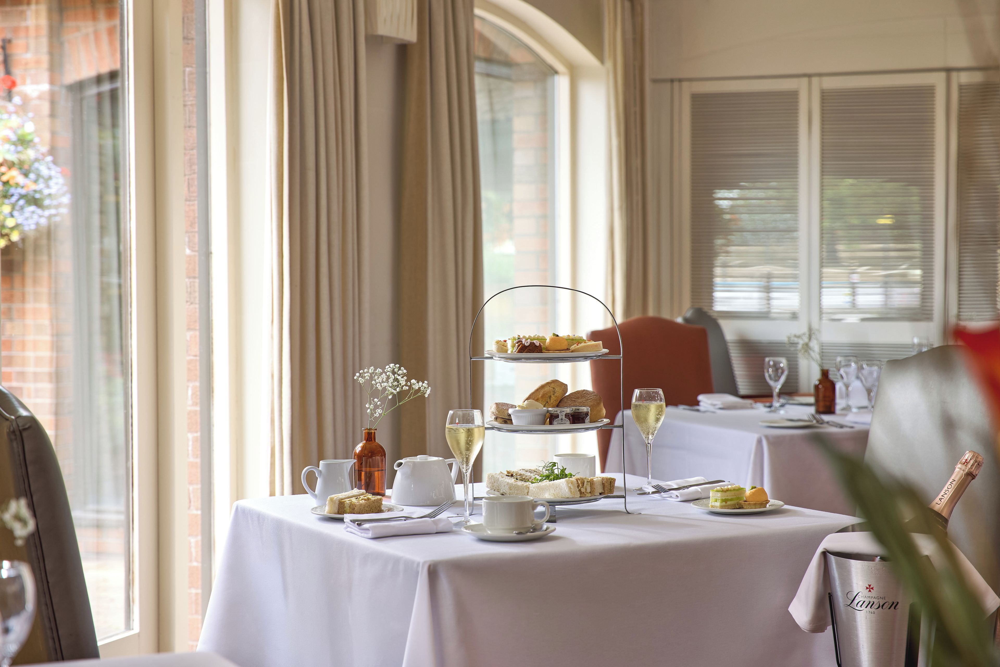 Un soporte de tres niveles con pasteles y un florero sobre una mesa, frente a una ventana con cortinas.