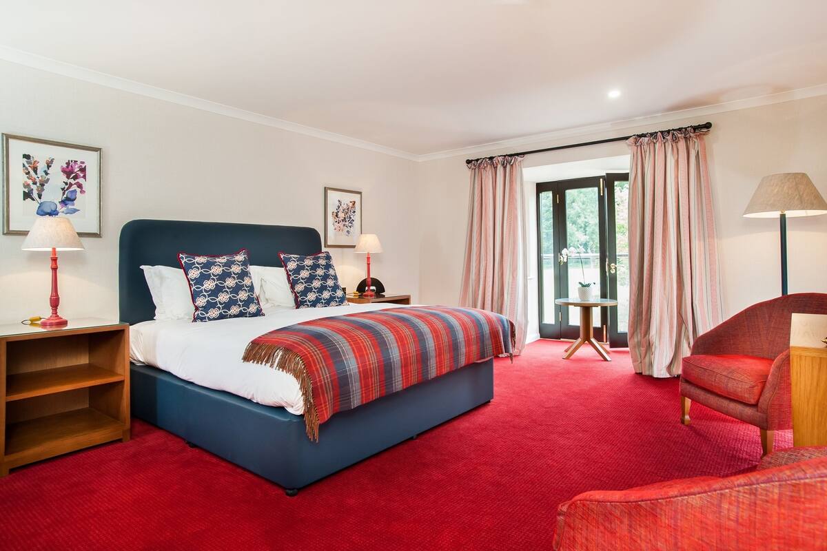 Una habitación de hotel con una cama grande, una alfombra roja, un sillón rojo y dos mesitas de noche con lámparas.