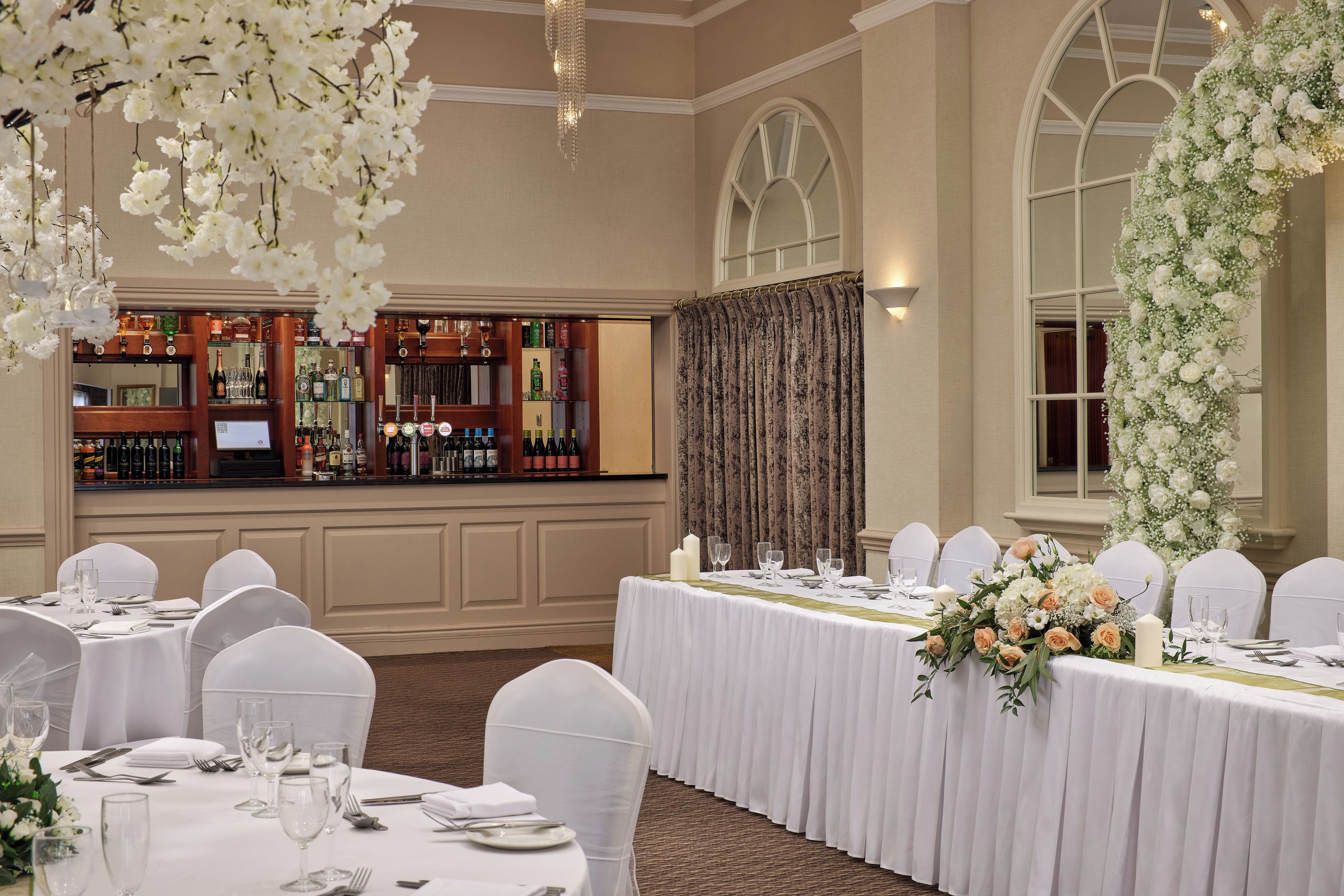 Un salón de banquetes con mesas redondas dispuestas para un evento formal, una zona de bar con diversas botellas de licor y decoraciones florales.