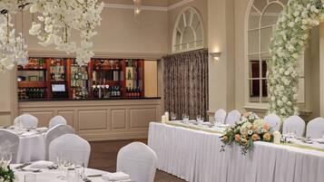 Un salón de banquetes con mesas redondas dispuestas para un evento formal, una zona de bar con diversas botellas de licor y decoraciones florales.