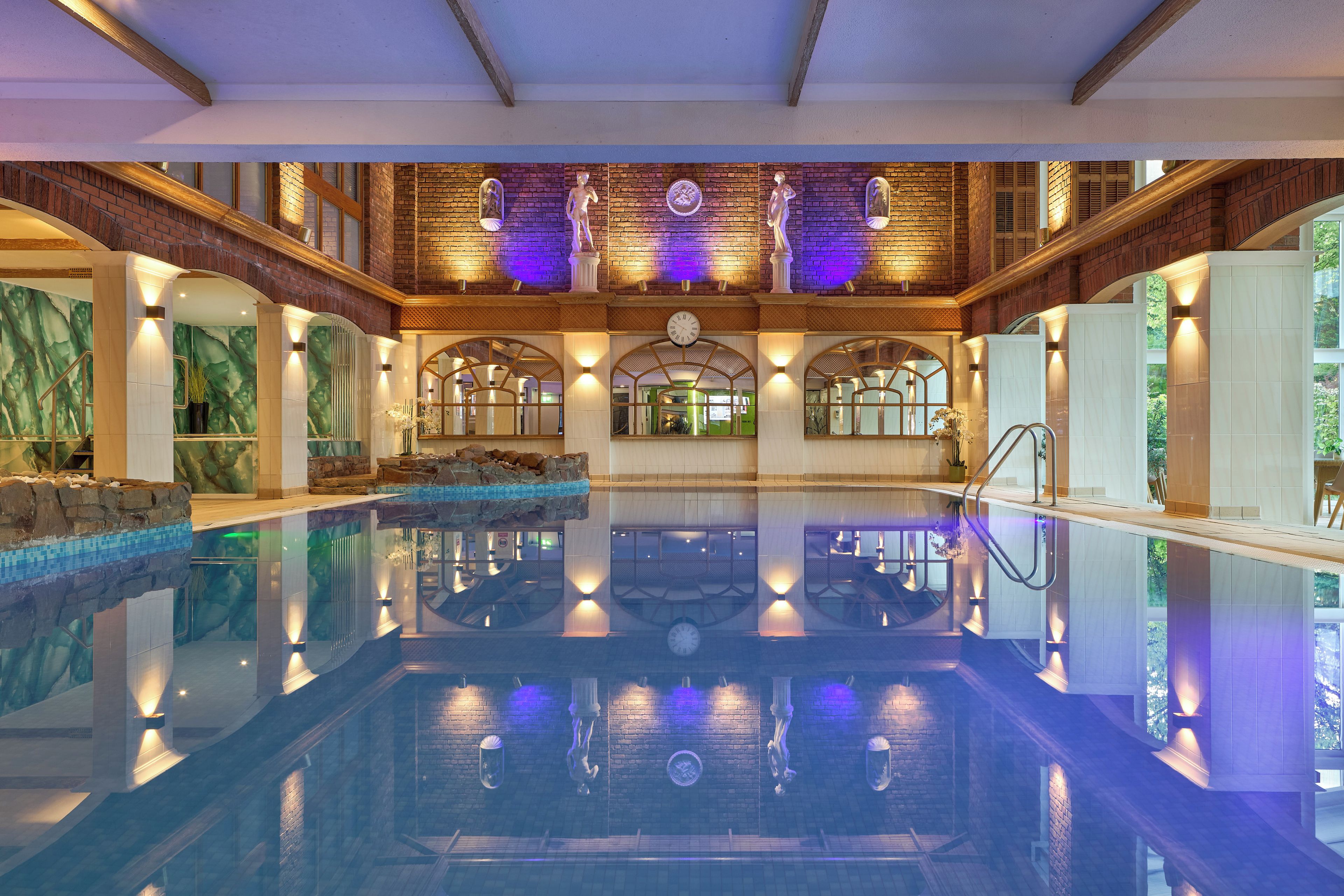 A large indoor swimming pool with a stone wall and arched windows, surrounded by a high ceiling with exposed beams.