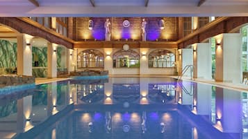 A large indoor swimming pool with a stone wall and arched windows, surrounded by a high ceiling with exposed beams.