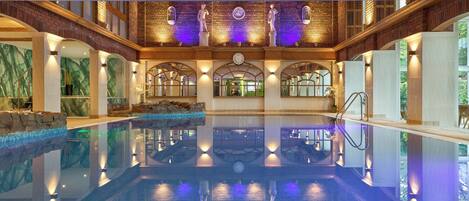 A large indoor swimming pool with a stone wall and arched windows, surrounded by a high ceiling with exposed beams.
