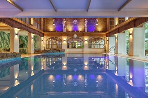 A large indoor swimming pool with a stone wall and arched windows, surrounded by a high ceiling with exposed beams.
