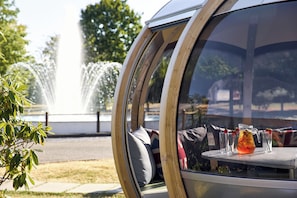 Un espace salon fermé par des parois de verre, avec vue sur une fontaine, une table avec des boissons et de la verdure.