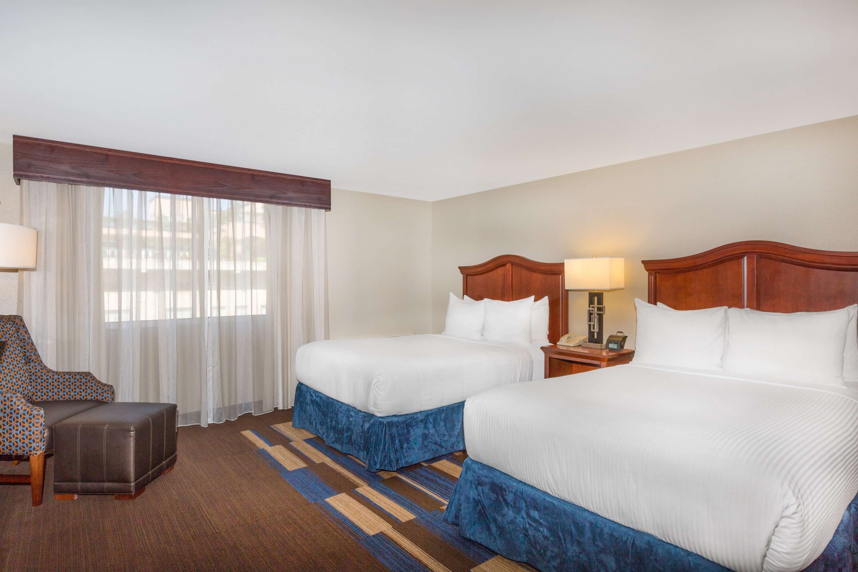 A hotel room with two beds, a desk, a chair, and a window with sheer curtains.