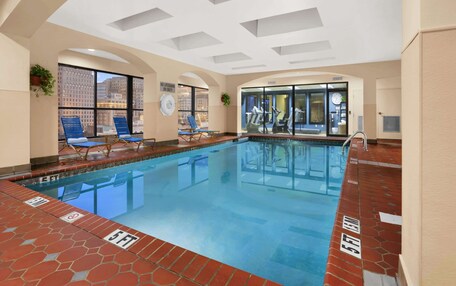 An indoor swimming pool with clear water, blue lounge chairs, and a view of the city through large windows.