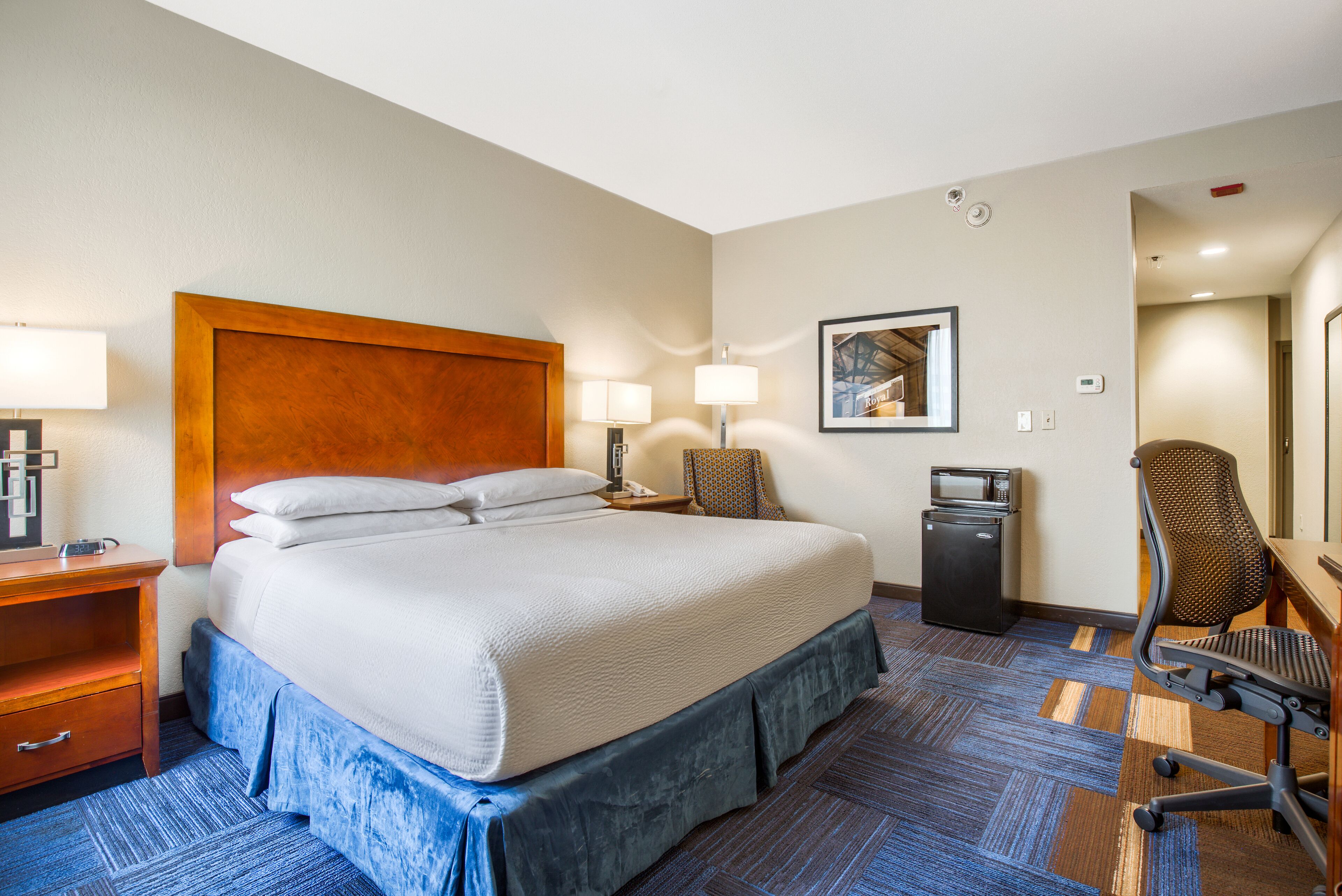 A hotel room with a large bed, two bedside tables with lamps, a desk with a chair, a microwave, and a framed picture on the wall.