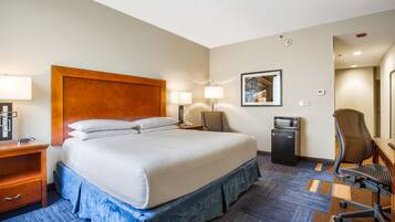 A hotel room with a large bed, two bedside tables with lamps, a desk with a chair, a microwave, and a framed picture on the wall.