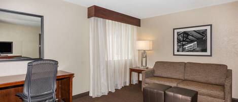 A hotel room with a desk, office chair, sofa, ottoman, and a large mirror.