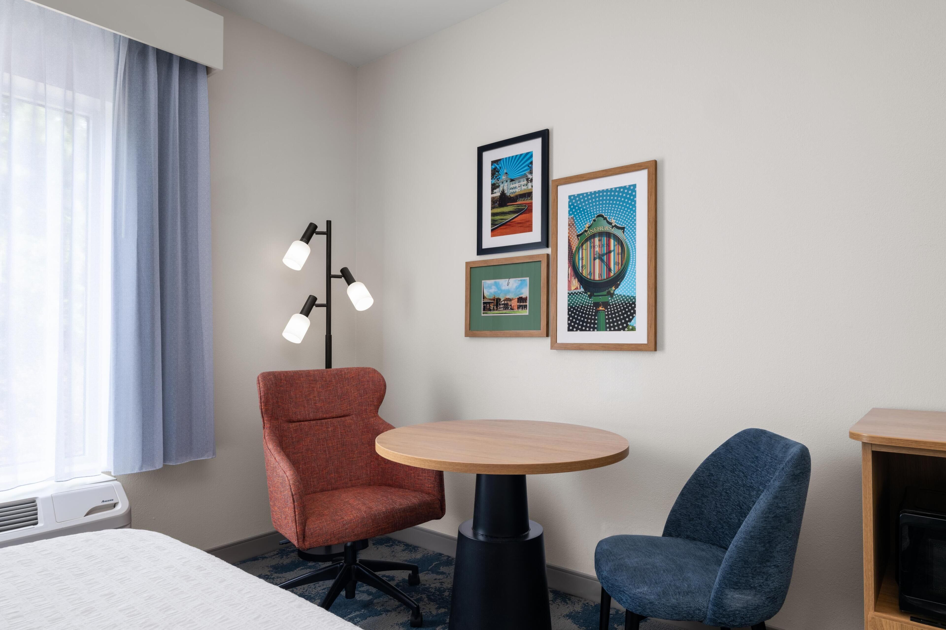 A hotel room with a bed, a round table, two chairs, a floor lamp, and three framed pictures on the wall.