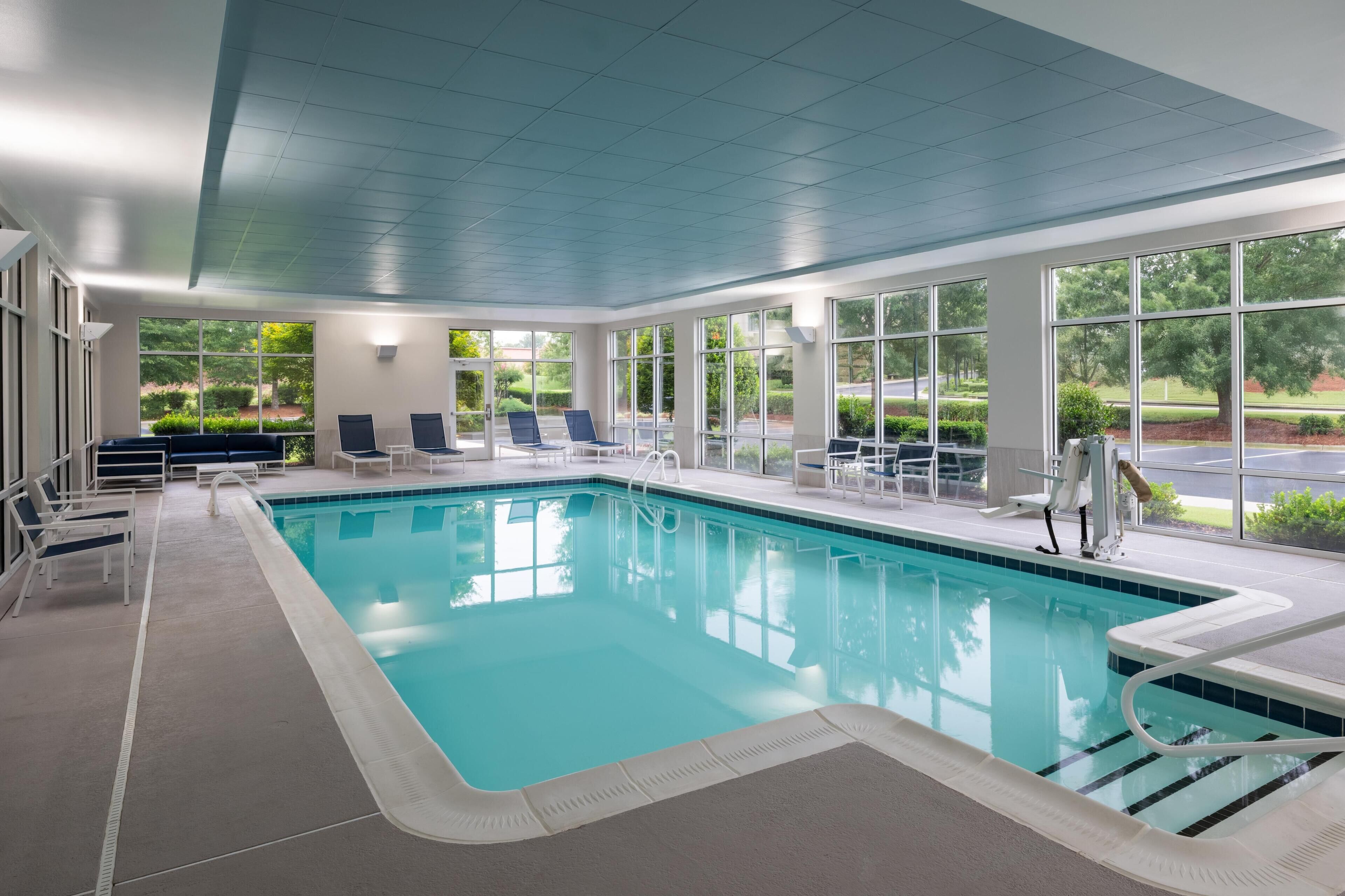 An indoor swimming pool with clear blue water, surrounded by white lounge chairs and a view of greenery outside.
