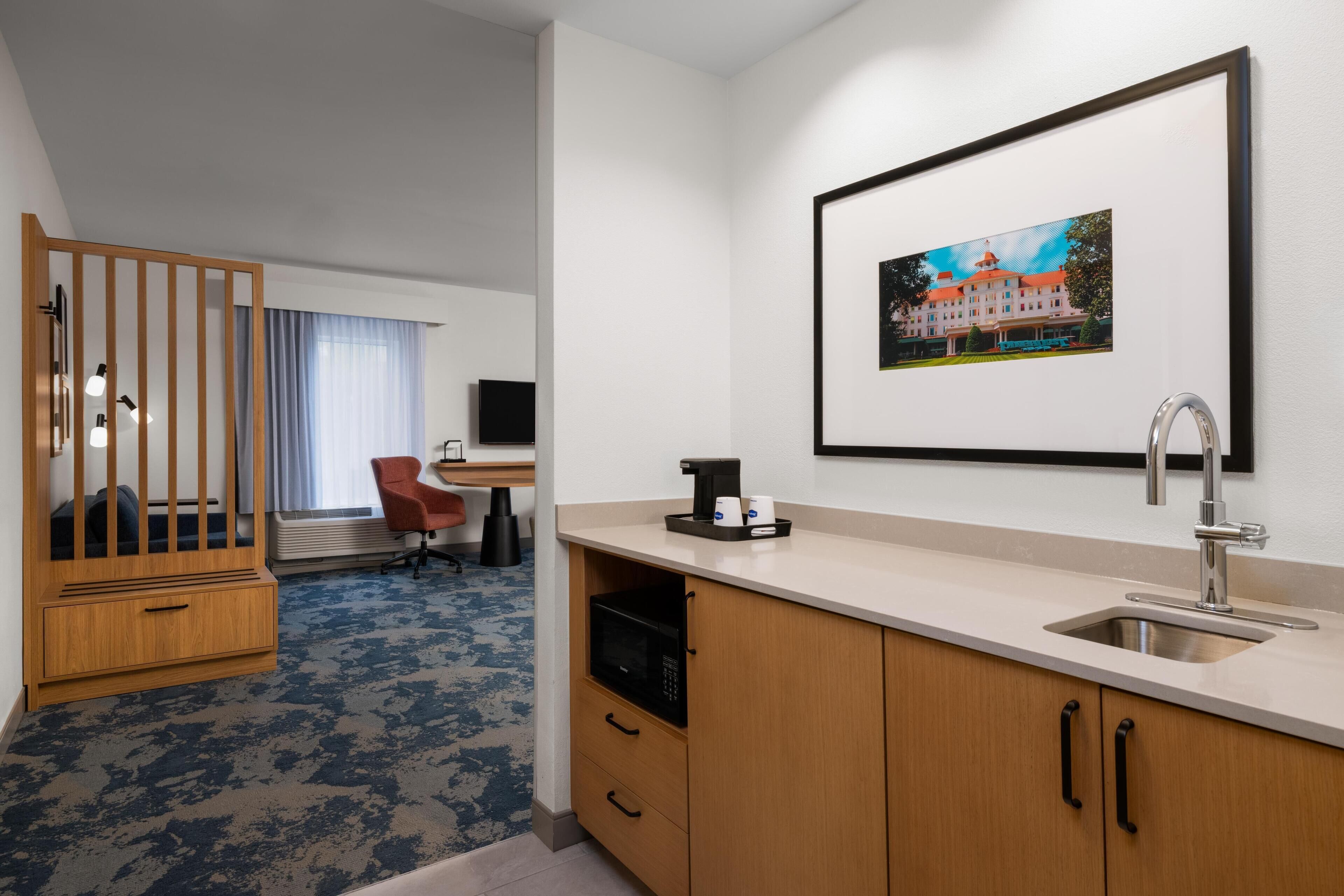 A hotel room with a kitchenette, a dining area, a TV, a bed, and a large window with curtains.