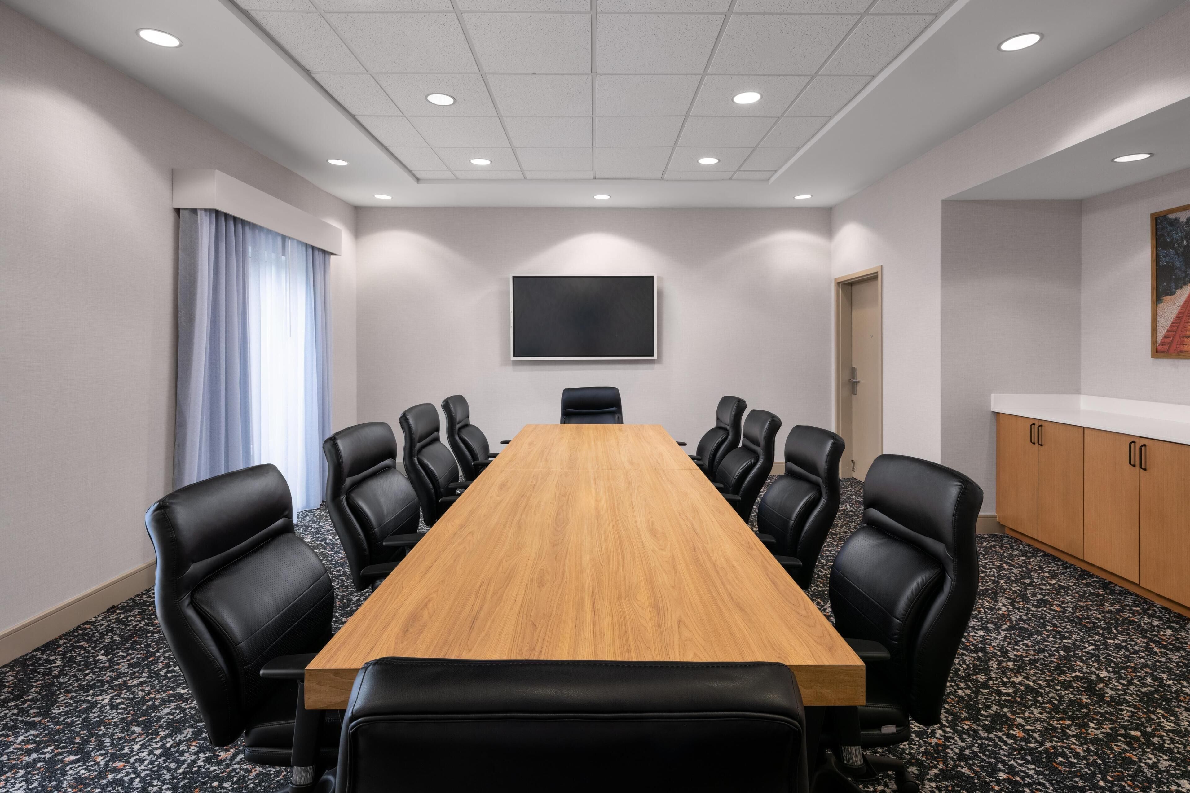 Une salle de conférence avec une longue table en bois, des fauteuils en cuir noir, un téléviseur à écran plat accroché au mur et une porte sur le côté droit.