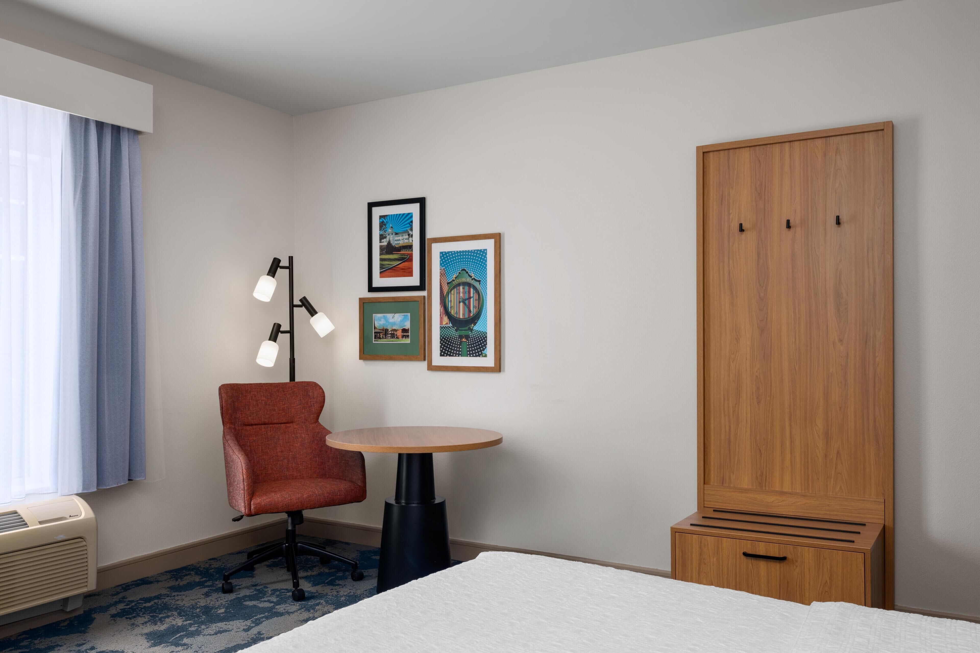 A hotel room with a bed, a red chair, a small table, a wardrobe, and three framed pictures on the wall.