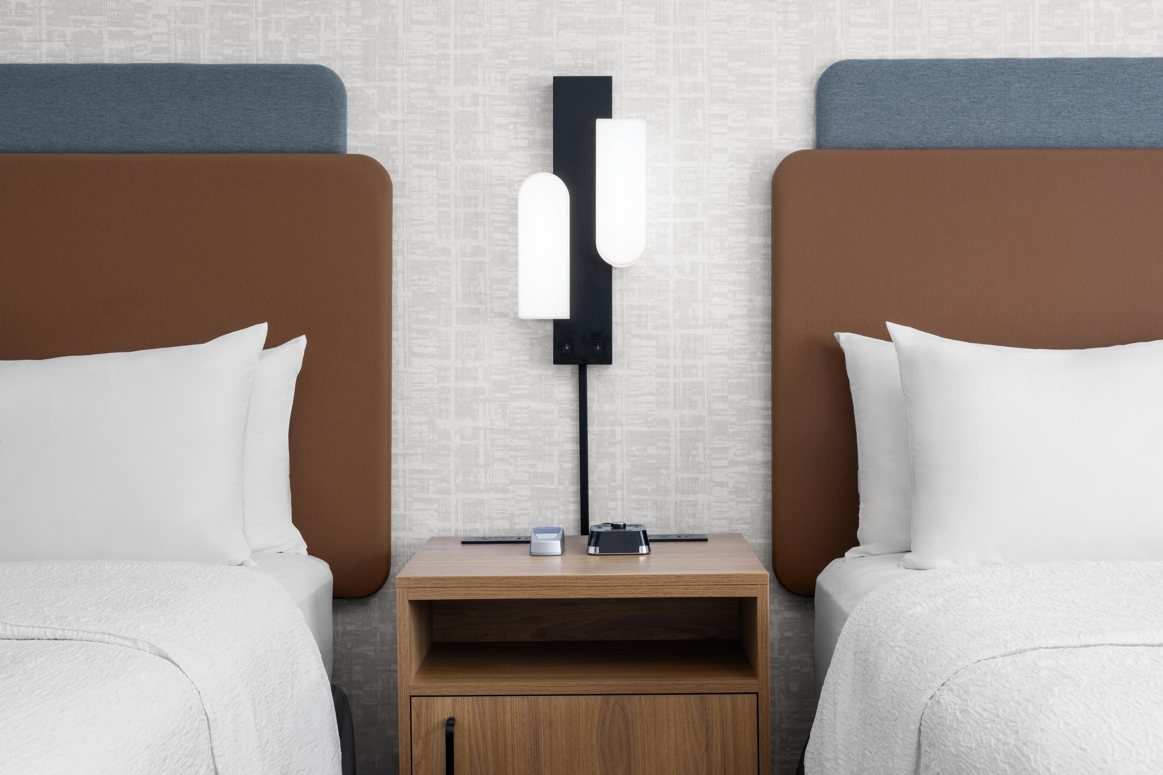 Quarto de hotel com duas camas, uma mesa de cabeceira com abajur e um pequeno armário de madeira.