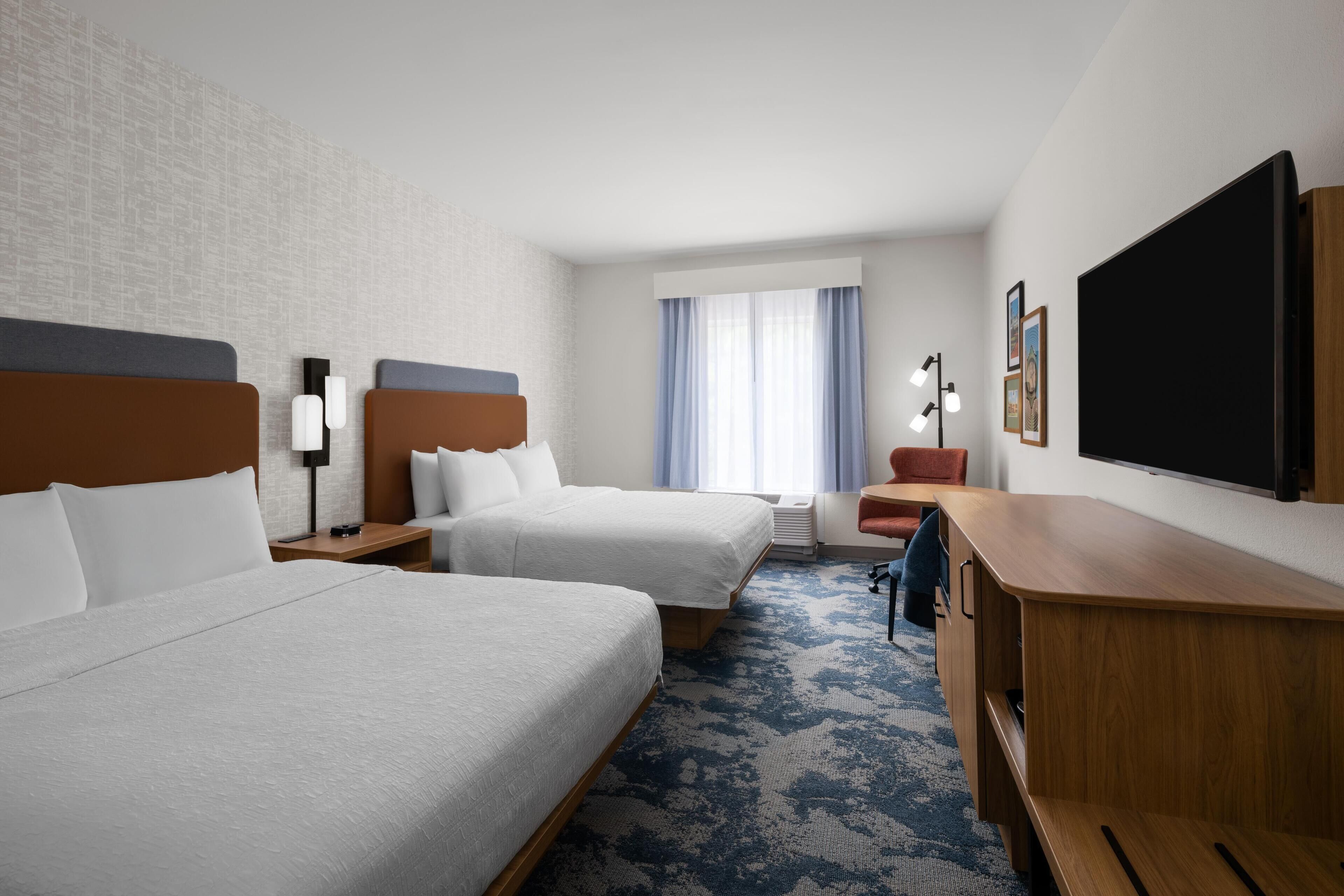 A hotel room with two beds, a desk, a chair, a TV, and a window with curtains.