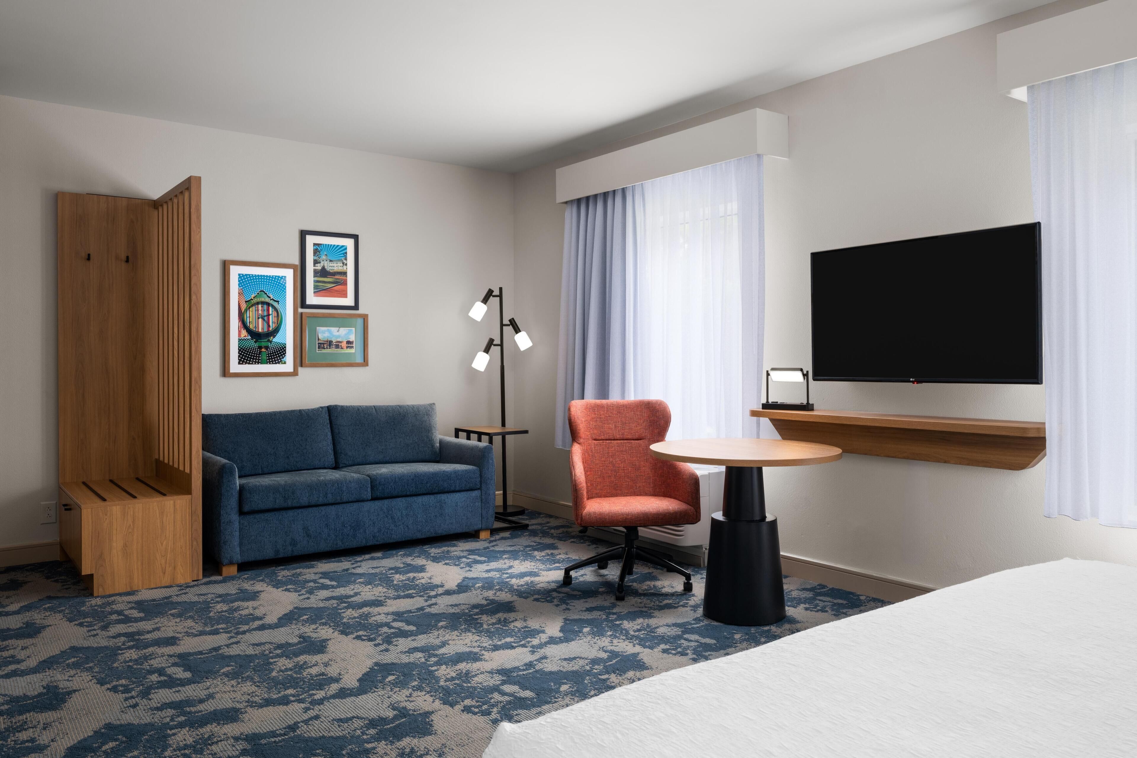 A hotel room with a blue sofa, a wooden cabinet, a round table, a TV, and artwork on the wall.