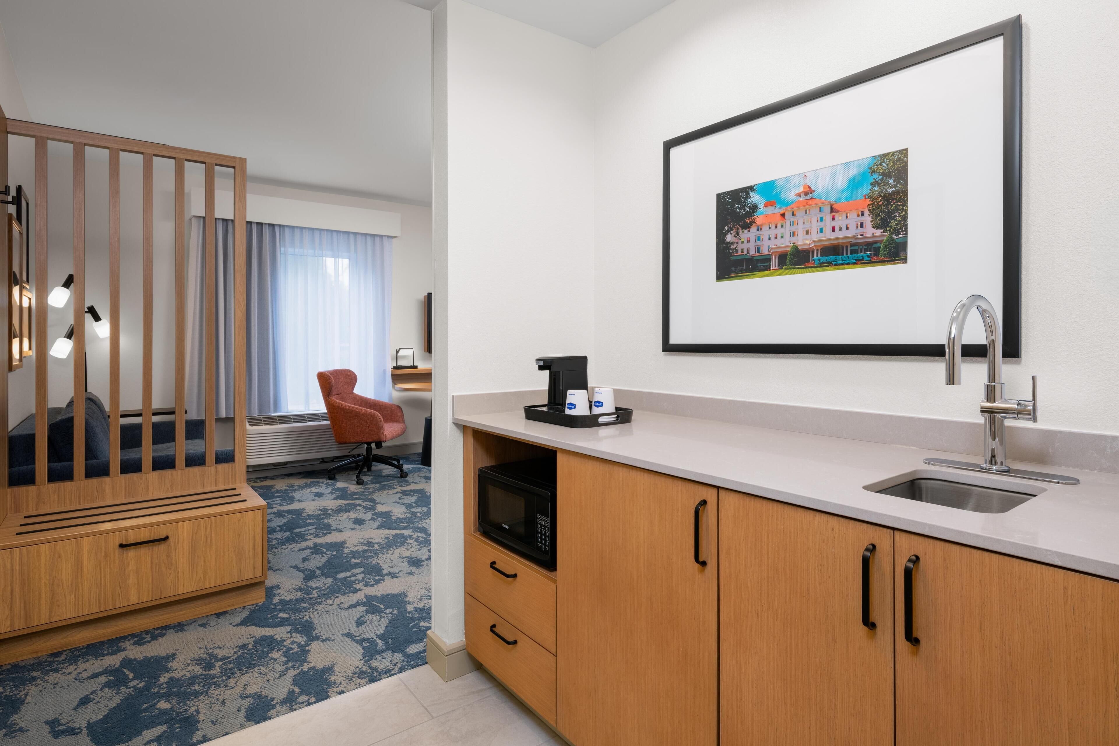 Una habitación de hotel con kitchenette, un mueble de madera, un microondas, un lavamanos, un cuadro en la pared, una ventana con cortinas y una alfombra azul.