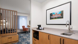 A hotel room with a kitchenette, a wooden cabinet, a microwave, a sink, a framed picture on the wall, a window with curtains, and a blue carpet.