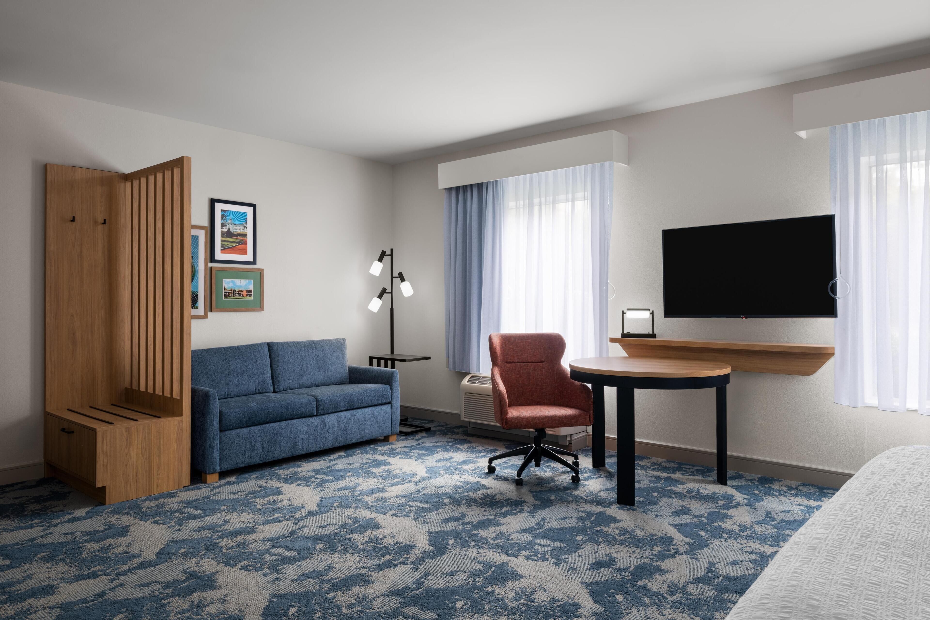 A hotel room with a blue sofa, a red chair, a small table, a TV mounted on the wall, and a large window with curtains.