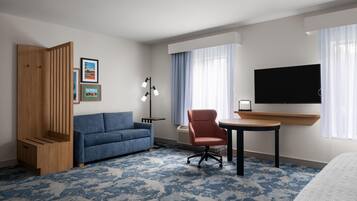 A hotel room with a blue sofa, a red chair, a small table, a TV mounted on the wall, and a large window with curtains.