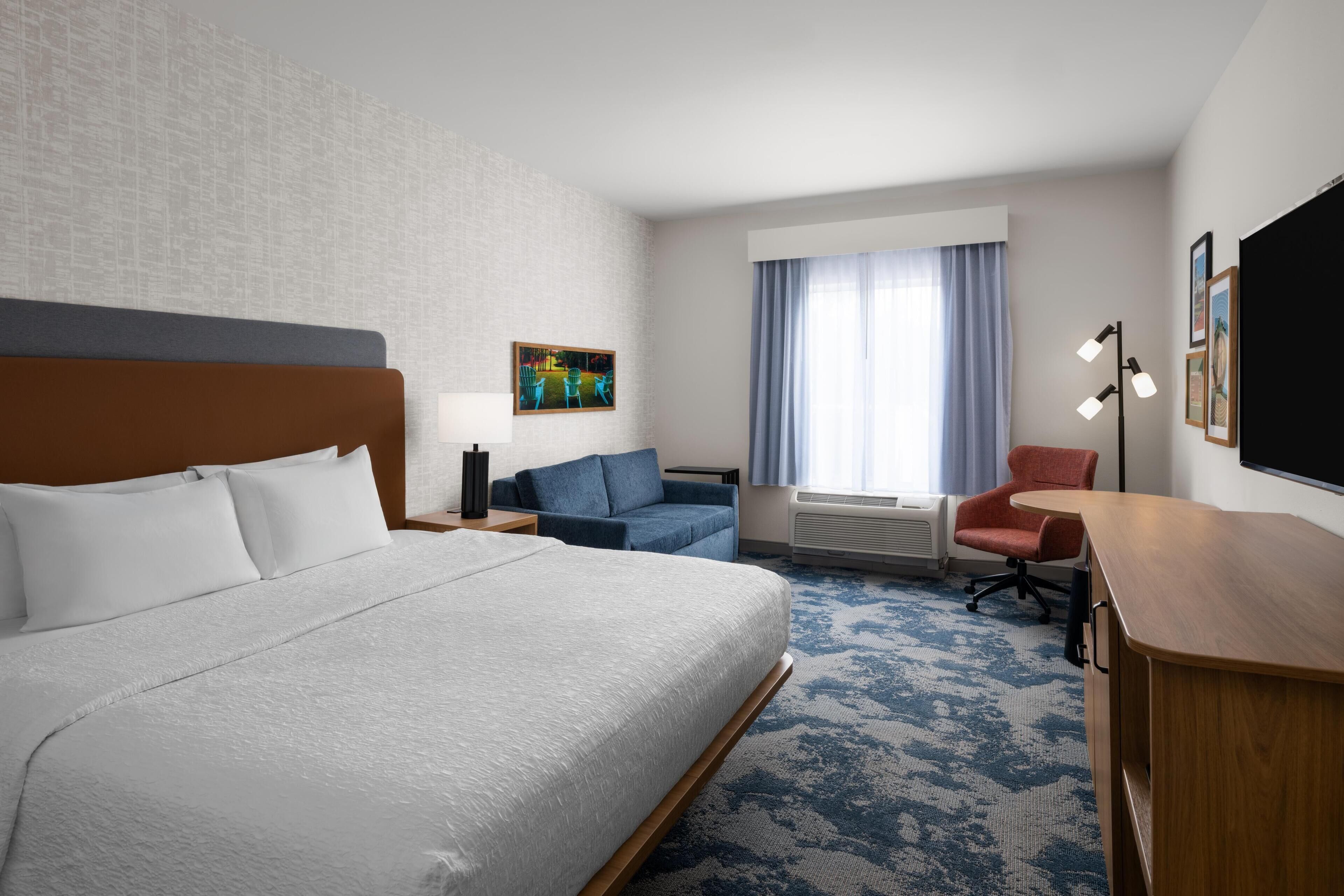 A hotel room with a large bed, a desk, a chair, a sofa, a lamp, and a TV.