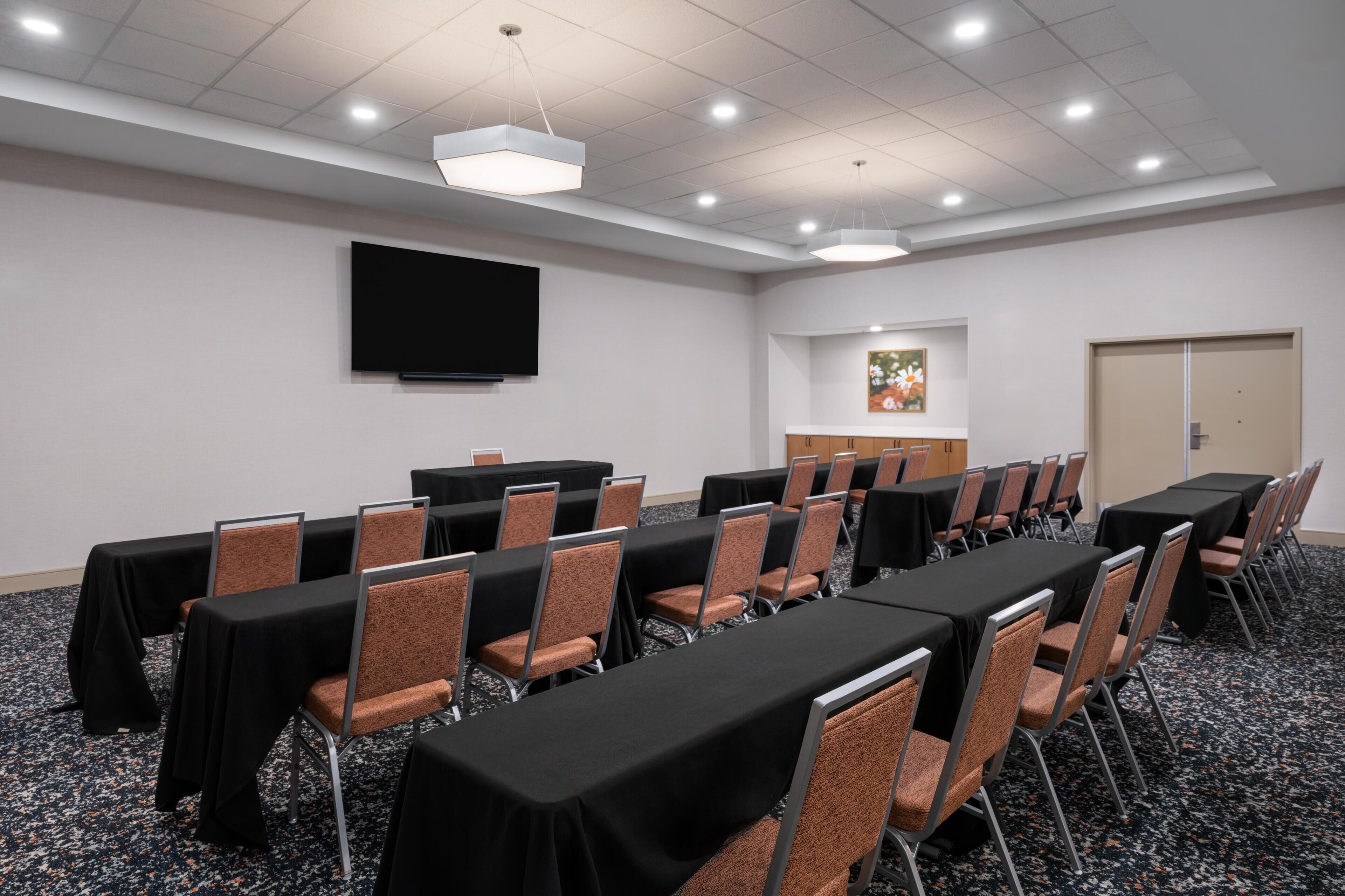 A conference room with rows of tables and chairs, a flat-screen TV, and a door.