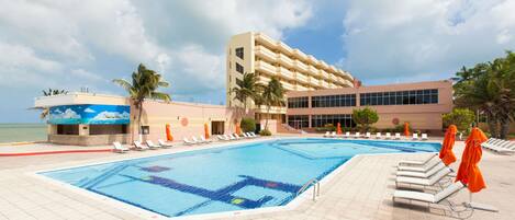 A swimming pool area with lounge chairs and umbrellas, a building with multiple floors, and a view of the ocean.