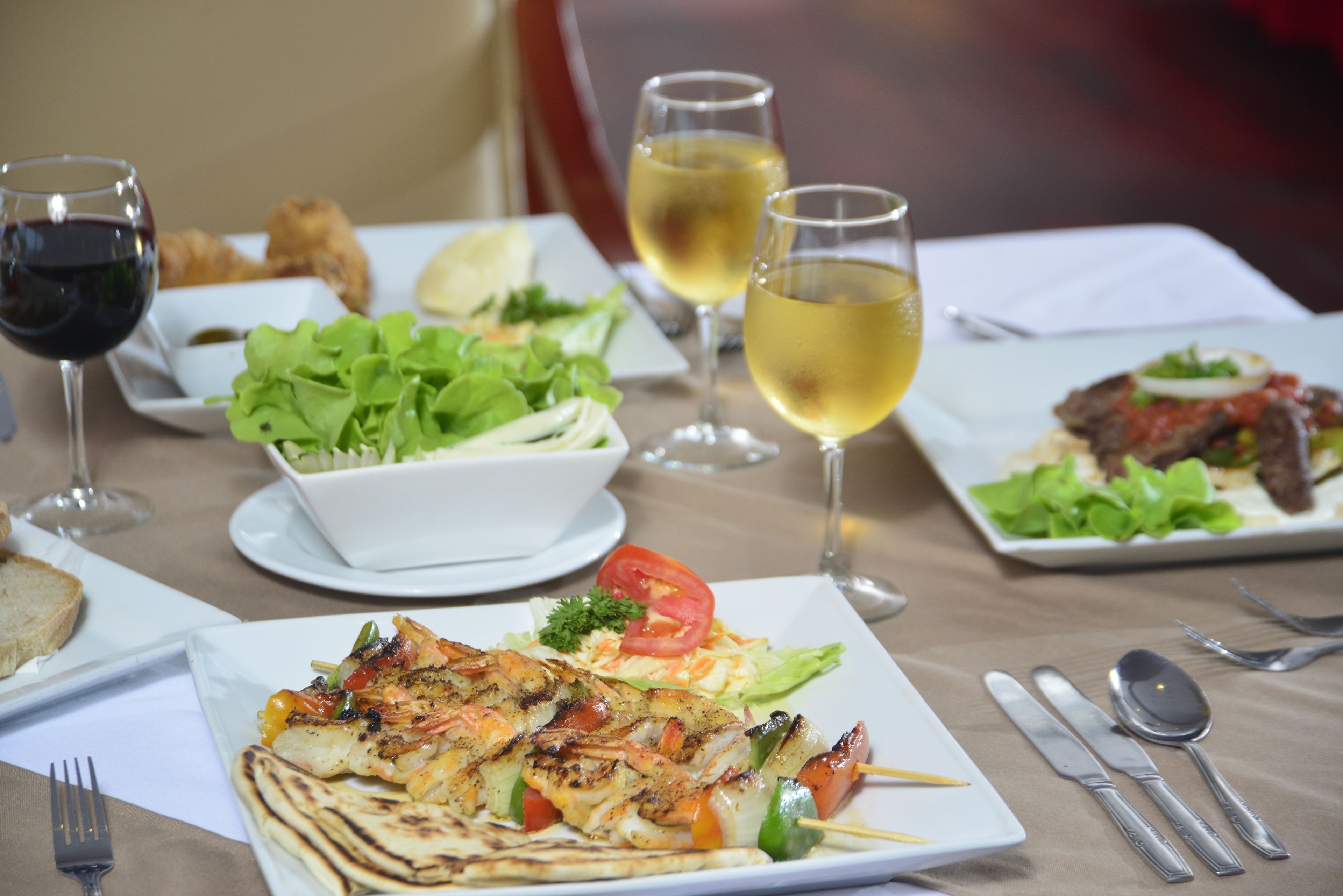 Una mesa de comedor con diversos platos, incluyendo mariscos a la parrilla, ensaladas y pan, junto con copas de vino y cubiertos.