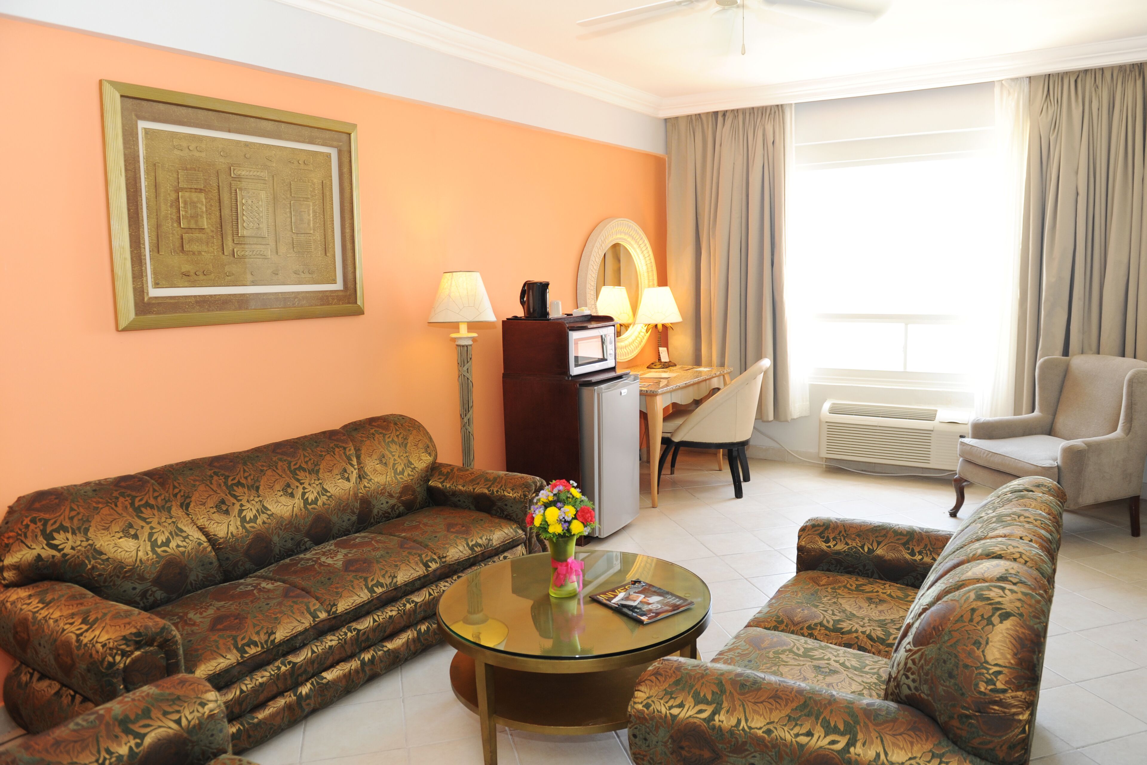 Habitación de hotel con sofá, sillón y una pequeña mesa con un jarrón de flores. Hay un escritorio con lámpara y televisor.