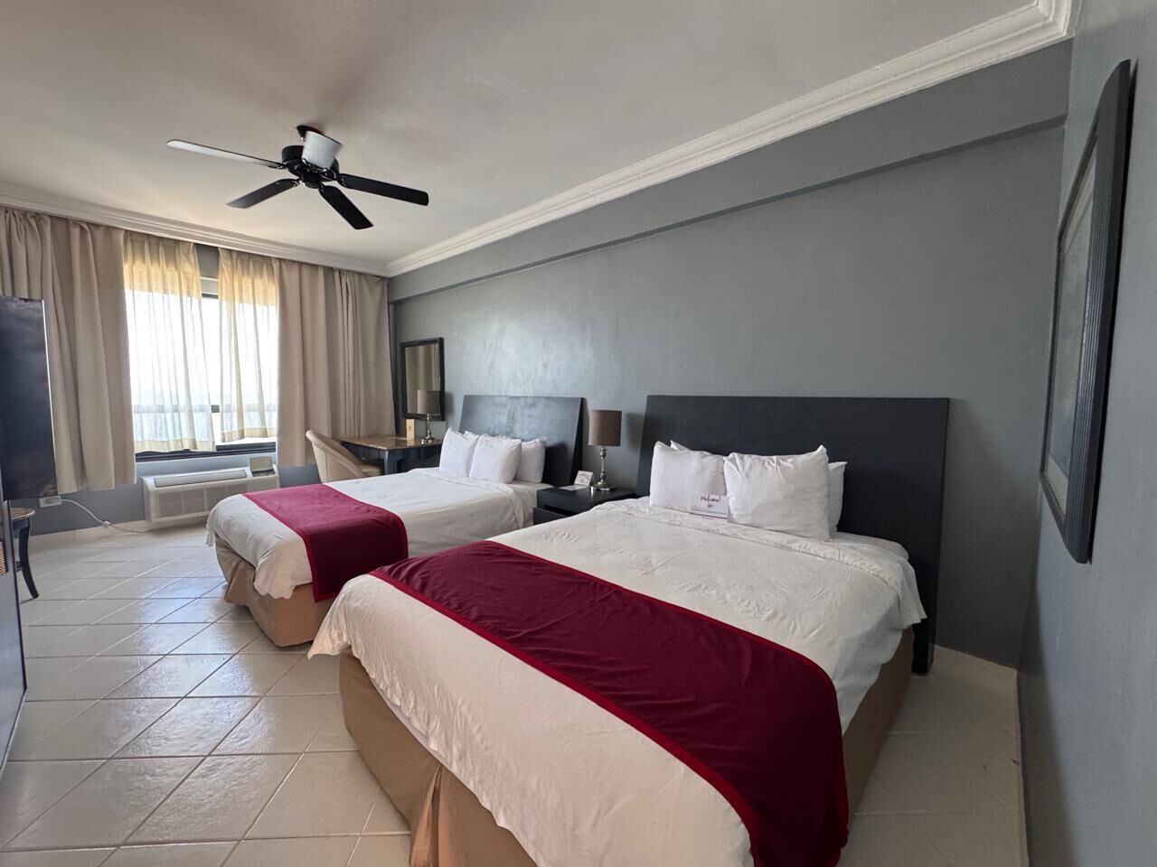 Habitación de hotel con dos camas, ventilador de techo, ventana con cortinas y una televisión de pantalla plana.