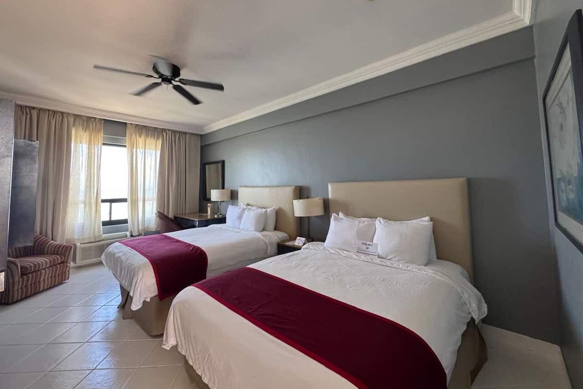 Een hotelkamer met twee bedden, een plafondventilator, een stoel en een groot raam met gordijnen.