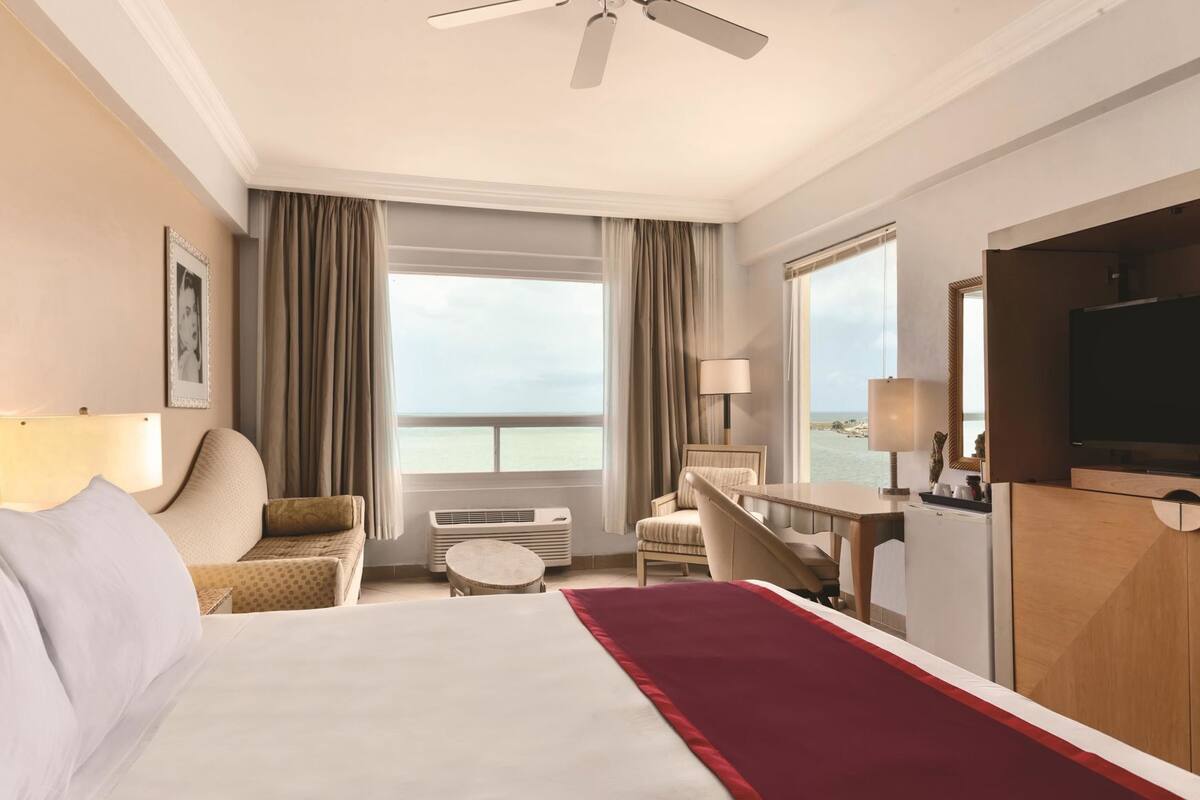 Een hotelkamer met een bed, bureau, stoel, televisie, raam met uitzicht op zee, en een plafondventilator.