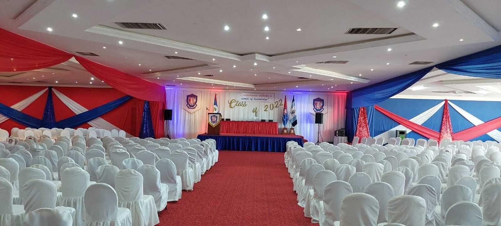 Une salle de banquet avec des rangées de chaises blanches, une scène avec un pupitre et des rideaux rouges et bleus.