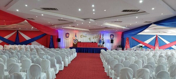 A banquet hall with rows of white chairs, a stage with a podium, and draped red and blue curtains.
