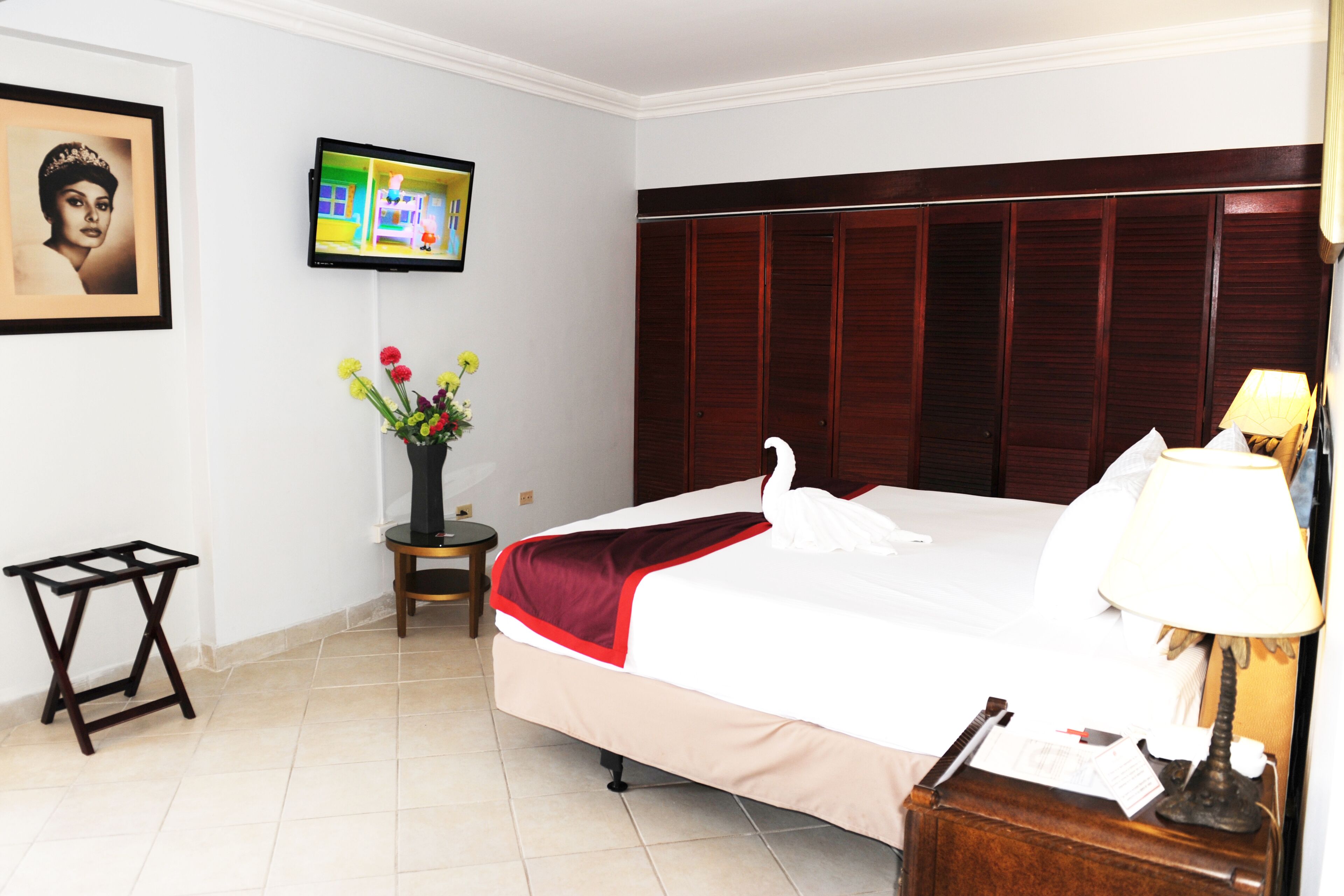 Habitación de hotel con una cama grande, un televisor montado en la pared, una mesita con un jarrón de flores y una lámpara en la mesita de noche.