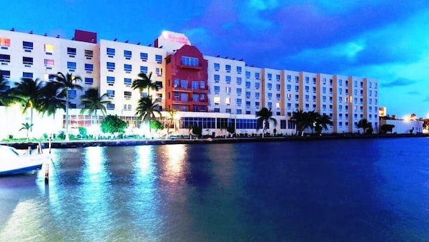 Un hotel con varios pisos, vista al agua y palmeras al frente.