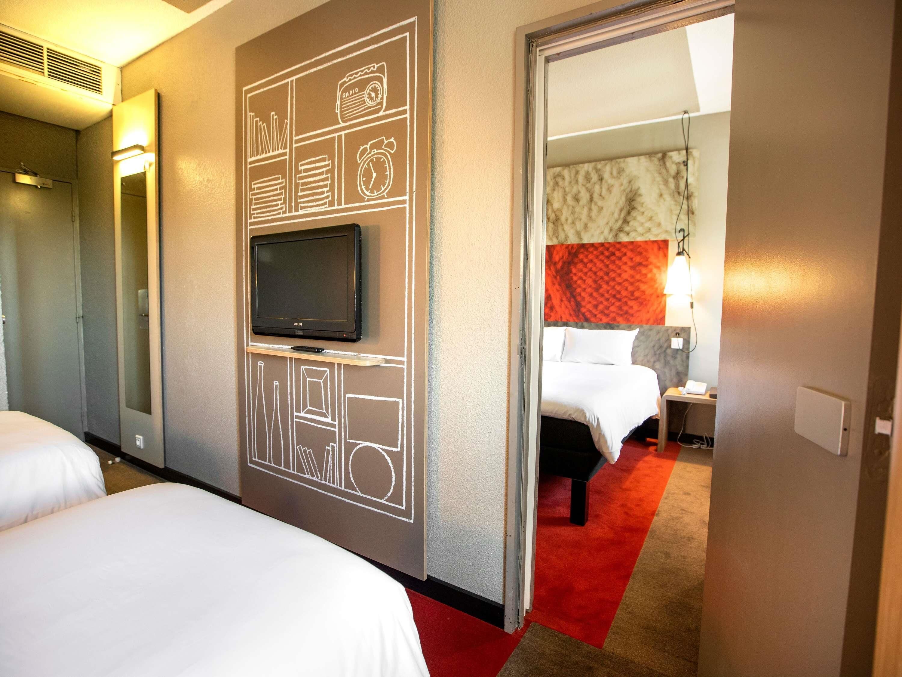 A hotel room with a large flat-screen TV mounted on the wall, a bed with white linens, and a red carpeted floor.