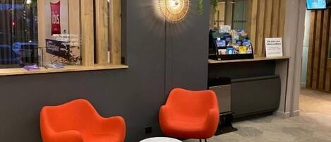 A reception area with two orange chairs, a small round table, a hanging light fixture, and a wooden counter with a sign.