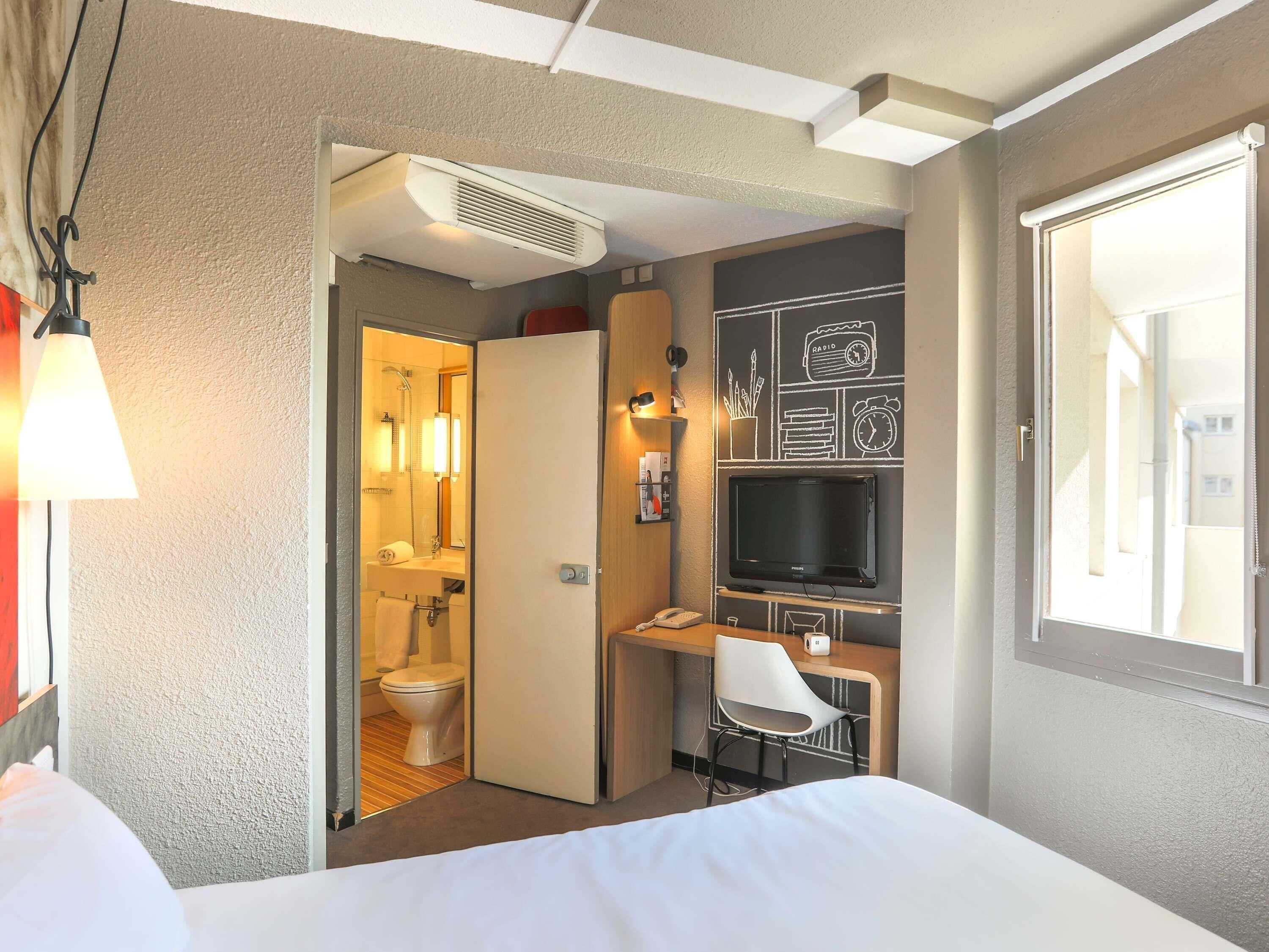 Habitación de hotel con cama, escritorio, televisor y baño visible a través de una puerta abierta.