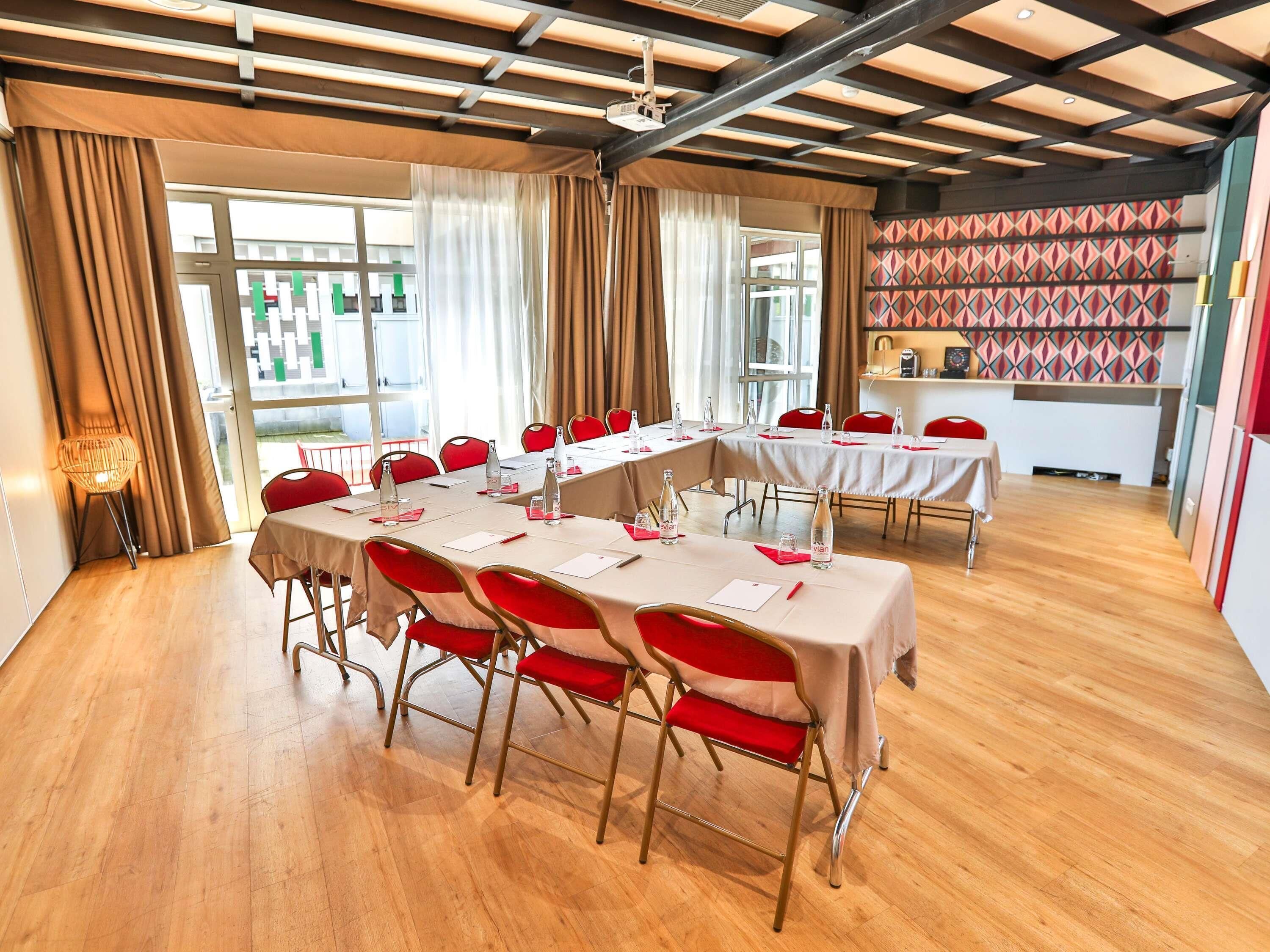 A conference room with multiple tables, red chairs, and a kitchen area visible through the windows.