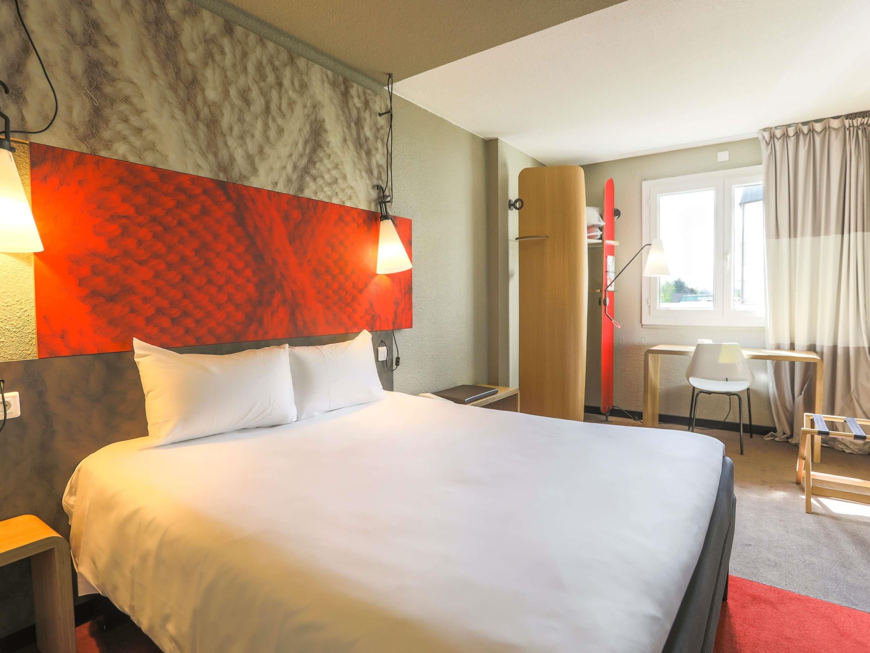 Een hotelkamer met een bed, bedlampen, een bureau, een stoel en een raam met gordijnen.