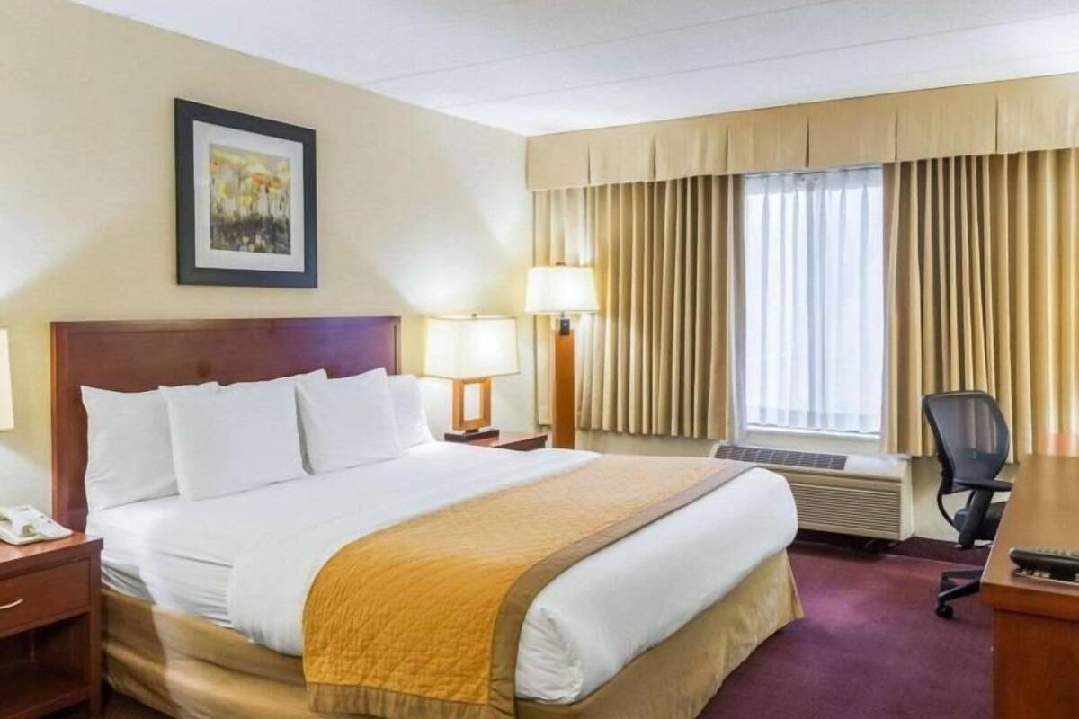 A hotel room with a bed, two bedside lamps, a desk with a chair, a window with curtains, and a painting on the wall.