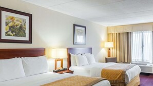 Hotellihuone, jossa on kaksi sänkyä, puinen pääty, yöpöytä lampun kanssa, kehystetty kukkamaalaus, ikkuna verhoilla ja ilmastointilaite.