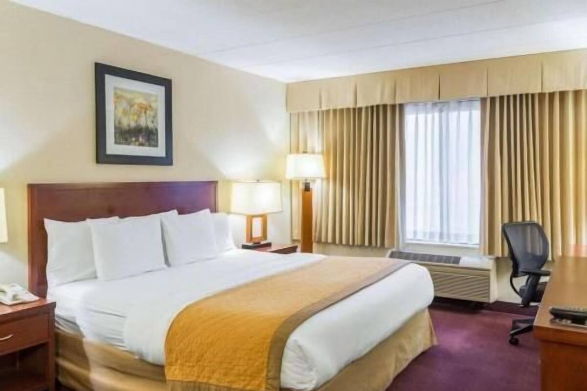 A hotel room with a bed, two bedside lamps, a desk with a chair, a window with curtains, and a painting on the wall.