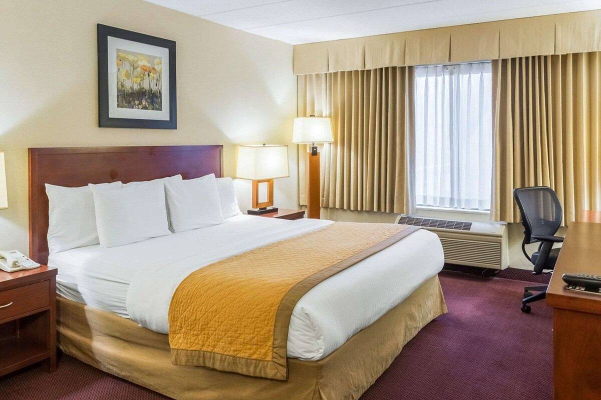 A hotel room with a large bed, two bedside lamps, a desk with a chair, a window with curtains, and a framed picture on the wall.