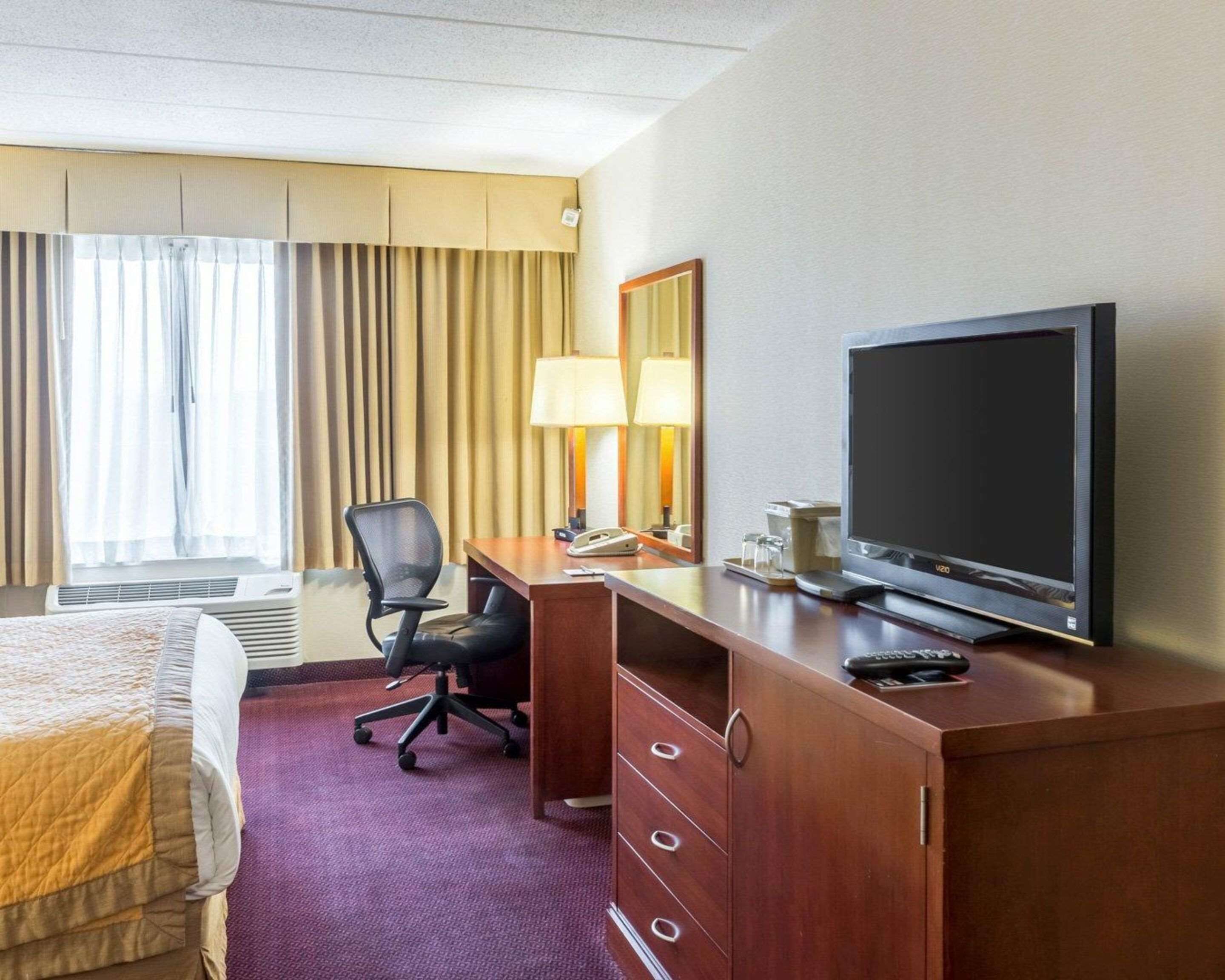 Une chambre d’hôtel avec un lit, un bureau avec une télévision, une chaise, une lampe et une fenêtre avec des rideaux.