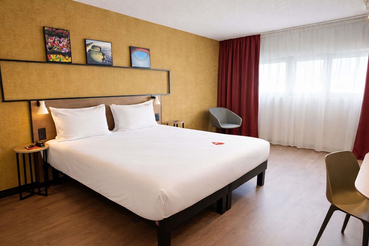 A hotel room with a large bed, a bedside table with a lamp, a chair, and artwork on the wall.