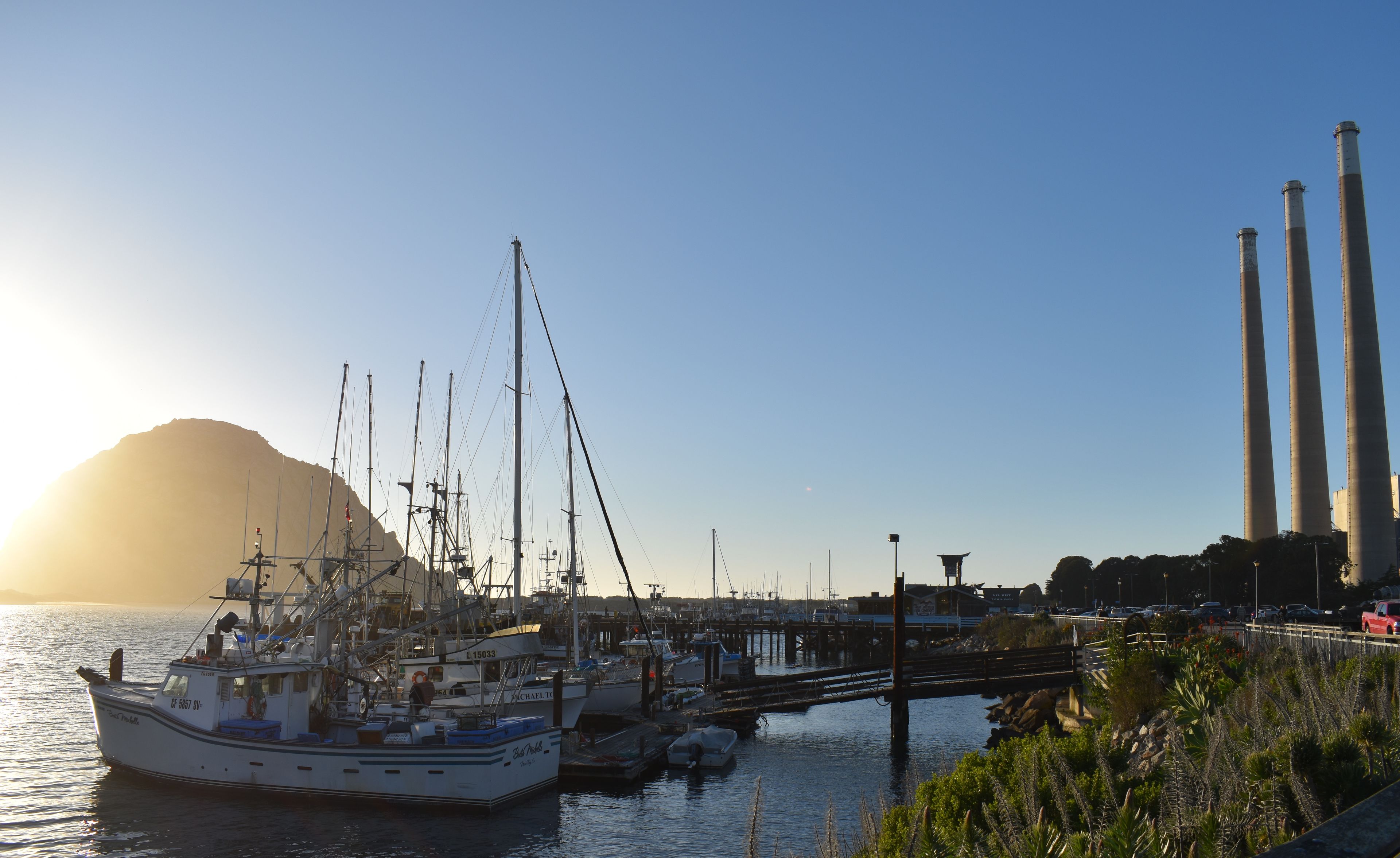 A marina with boats docked, a bridge, and industrial chimneys in the background.